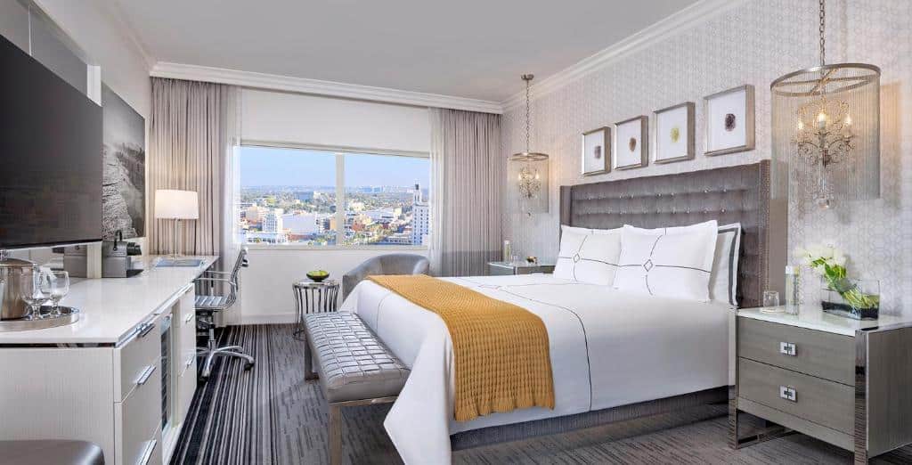 Quarto no Huntley Santa Monica Beach com trinta e três metros quadrados, com uma cama de casal, uma janela com vista para cidade com cortinas, luminárias suspensas uma de cada lado da cama, uma mesinha de trabalho com uma cadeira giratória, uma televisão e o chão é de madeira, o local todo é decorado em cinza e os móveis tem detalhes em prata.