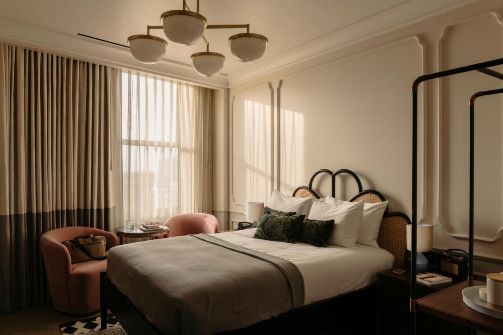 Um quarto iluminado pelo sol no The Hoxton, Downtown LA, com uma cama, duas cadeiras cor-de-rosa perto de uma janela e uma mesa de cabeceira com uma xícara e um pires, para representar hotéis em Los Angeles.