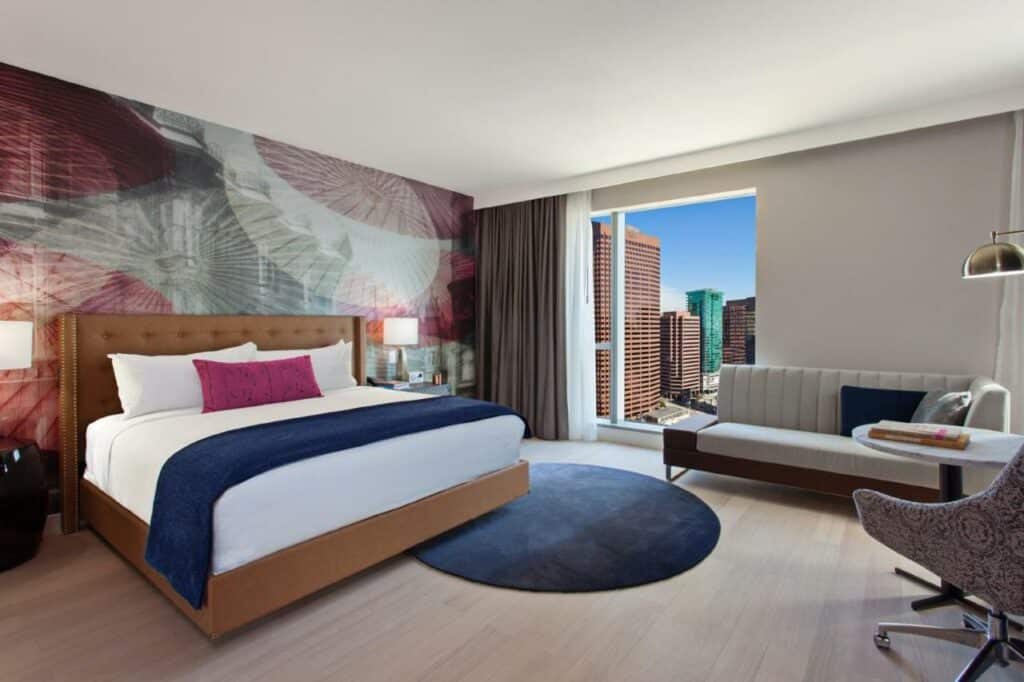 Quarto de quarenta e dois metros quadrados do Hotel Indigo - Los Angeles Downtown, an IHG Hotel com uma cama de casal, uma janela ampla com cortinas, um sofá cinza ao lado e uma poltrona também cinza, um tapete azul-marinho redondo perto da cama, tudo decorado em tons de cinza, azul e rosa escuro.