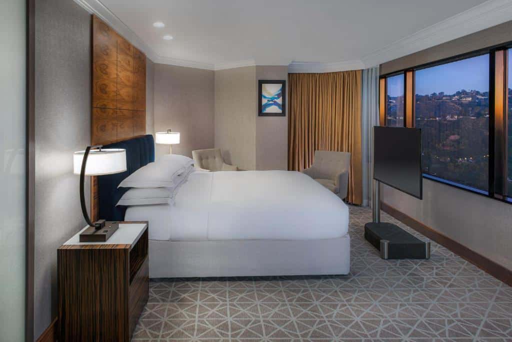 Quarto no Hilton Los Angeles-Universal City com chão de carpete com desenhos geométricos em branco com fundo bege, uma cama de casal de frente para uma janela ampla com vista para a cidade, além de uma televisão, há também duas poltronas beges e duas mesinhas de cabeceira de madeira com abajures, para representar hotéis em Los Angeles.