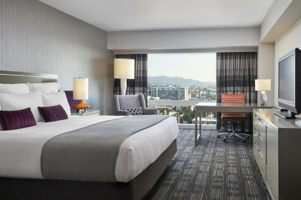 Quarto do Loews Hollywood Hotel com trinta e sete metros quadrados, um chão de carpete cinza com preto, uma janela panorâmica com cortinas, uma mesinha de escritório com uma cadeira giratório, do outro lado, perto da cama de casal, há uma poltrona e um abajur de chão, de frente para a cama, há uma televisão e uma cômoda de madeira, para representar hotéis em Los Angeles.