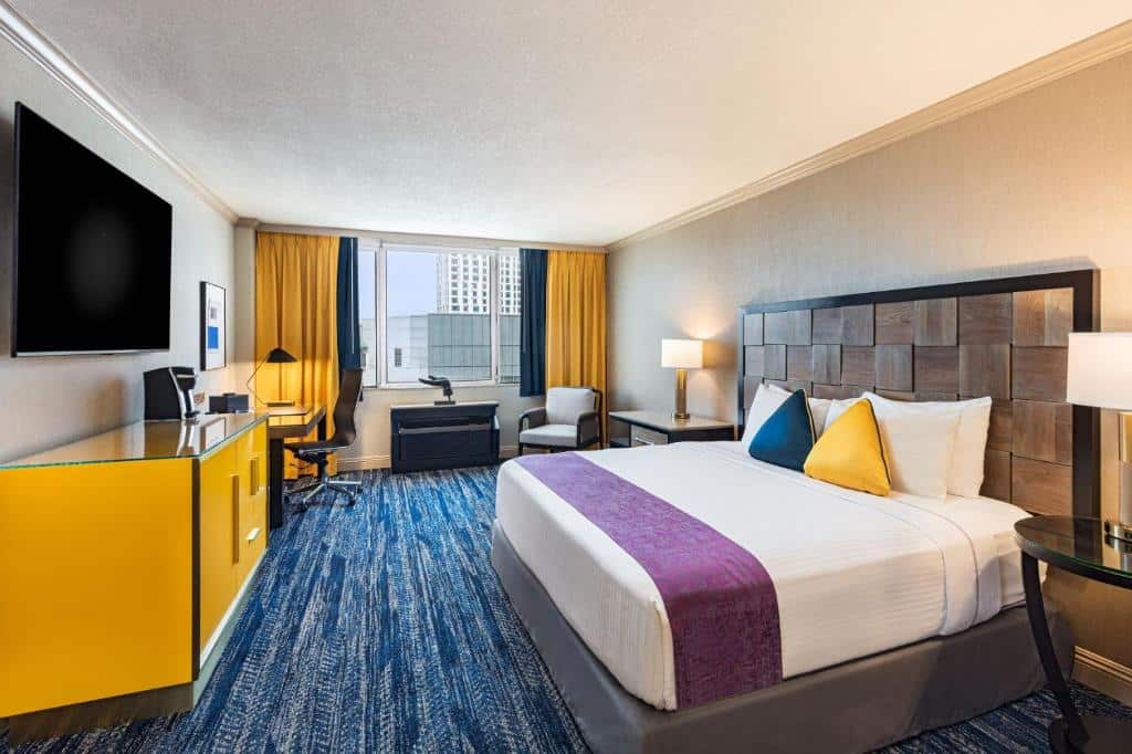 Quarto do E Central Hotel Downtown Los Angeles com vinte e nove metros quadrados, com um carpete em tons de azul-marinho e branco, uma janela ampla com cortinas azul-marinho e amarelo, do lado há uma poltrona estofada branca, depois uma pequena mesinha com um abajur ao lado de cama de casal, de frente para a cama tem uma cômoda amarela com uma televisão presa na parede, e mais ao lado, uma mesinha de escritório com uma cadeira giratória.