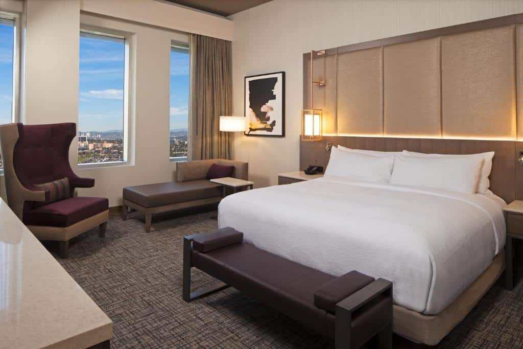 Quarto do H Hotel Los Angeles, Curio Collection By Hilton com trinta e cinco metros quadrados, três janelas compridas, uma poltrona estofada em vinho, um pequeno sofá de um lugar com um abajur do lado, uma cama de casal com luminárias suspensas do lado, o chão é de carpete cinza com preto, para representar hotéis em Los Angeles.