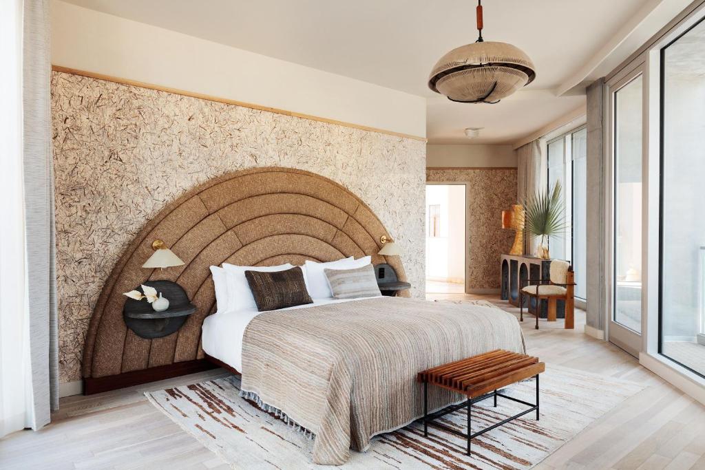 Quarto amplo no Santa Monica Proper Hotel, a Member of Design Hotels com oitenta e seis metros quadrados, com uma cama de casal, uma cabeceira arredondada rústica com luminárias presas nela, o chão é de madeira, e há uma porta de vidro que leva para outro ambiente, toda a decoração é em bege, marrom e detalhes em madeira.