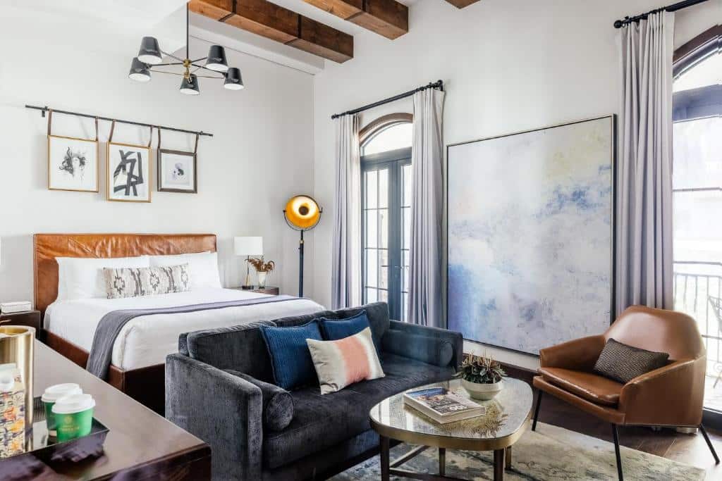 Quarto do Hotel Figueroa, Unbound Collection by Hyatt com trinta e dois metros quadrados, decorado em estilo contemporâneo com estilo vintage, há duas portas que levam para a sacada, do lado de dentro é possível ver uma pequena poltrona de couro marrom, uma mesinha de centro com livros em cima e uma sofá cinza com almofadas, para representar hotéis em Los Angeles.
