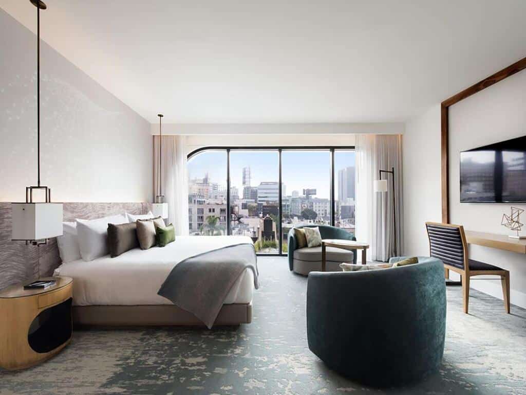 Quarto do Dream Hollywood. Uma cama de casal do lado esquerdo com um lustre e uma cômoda de cada lado. Do lado direito duas poltronas, uma mesa de trabalho e uma TV. No fundo uma parede de vidro.
