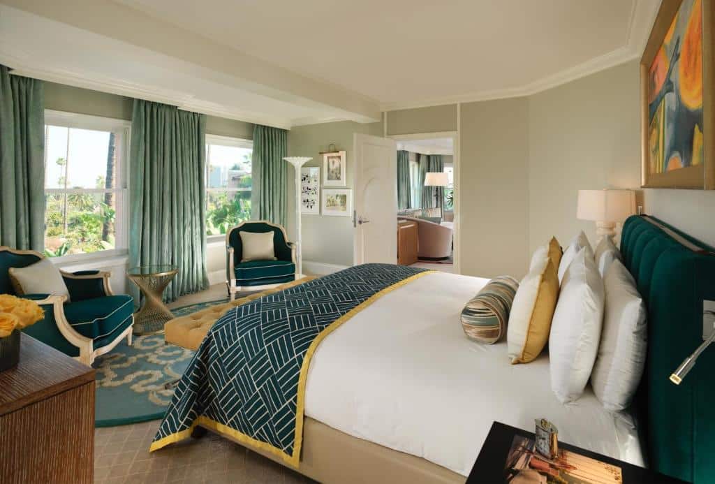 Quarto no The Beverly Hills Hotel - Dorchester Collection com setenta e dois metros quadrados, há duas janelas de frente para a cama de casal que está cheia de almofadas e travesseiros, há duas poltronas em direção das janelas, um tapete em tom de azul claro, o quarto é decorado em verde escuro e branco.