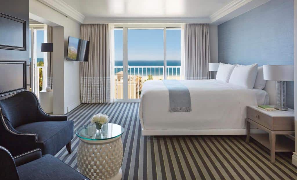 Quarto do Viceroy Santa Monica com cinquenta e seis metros quadrados, com uma cama de casal, uma sacada com vista direta para a praia, o chão é de carpete em listras cinza e azul, do lado esquerdo há duas poltronas azuis e uma televisão, para representar hotéis em Los Angeles.