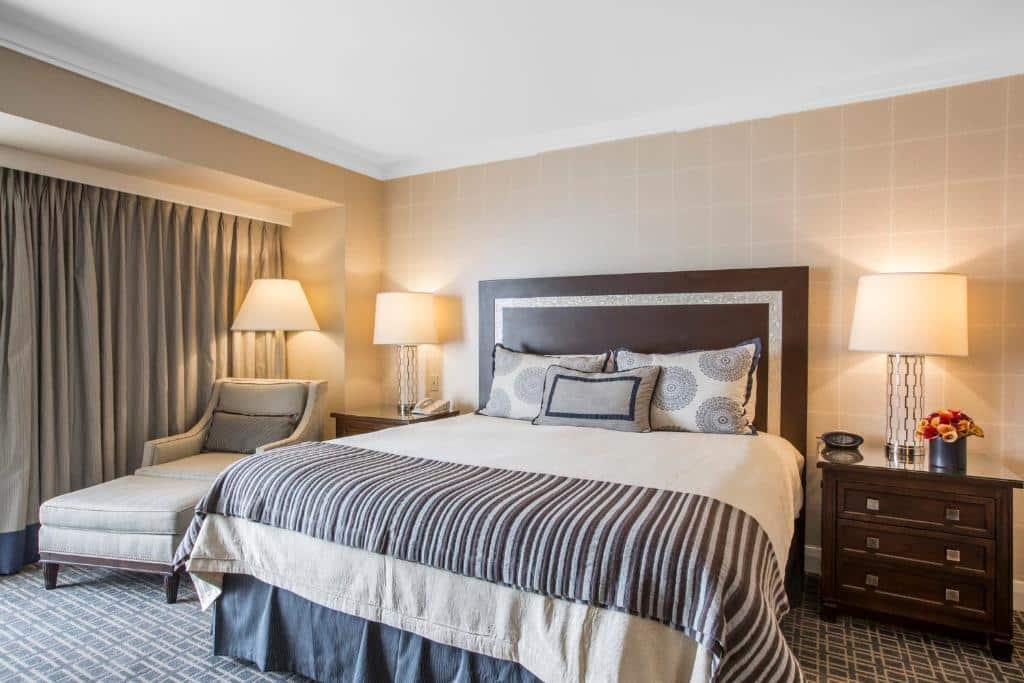 Quarto no Omni Los Angeles Hotel California Plaza com trinta e dois metros quadrados, uma cama de casal, uma pequena cômoda com um abajur em cima ao lado direito da cama, do lado esquerdo da cama há uma poltrona cinza, o chão é de carpete cinza com detalhes brancos.