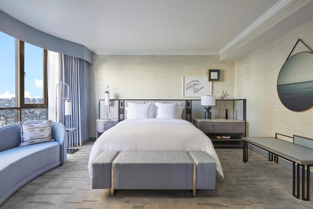 Quarto do Beverly Wilshire, A Four Seasons Hotel com sessenta e sete metros quadrados, do lado esquerod há uma janela cm cortinas cinzas, um sofá cinza com dois lugares, no centro da foto a cama de casal, do lado direito uma cômoda com duas gavetas e um abajur, o local inteiro é decorado em tons de cinza e branco, para representar hotéis em Los Angeles.