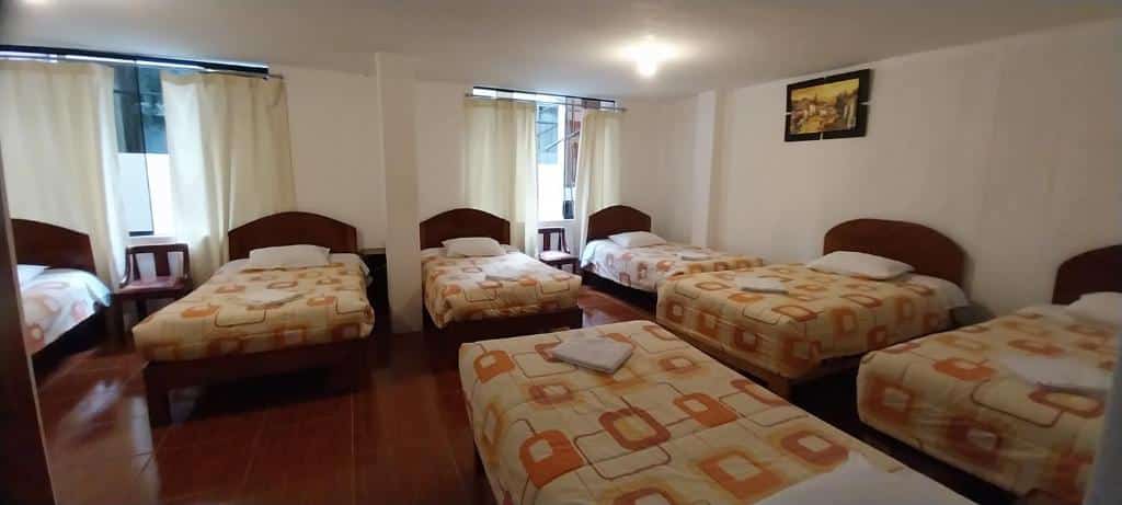 quarto com sete camas de solteiro dispostas em um mesmo ambiente. É um espaço simples com lençóis coloridos e duas janelas cobertas por uma cortina em cada lado do quarto.