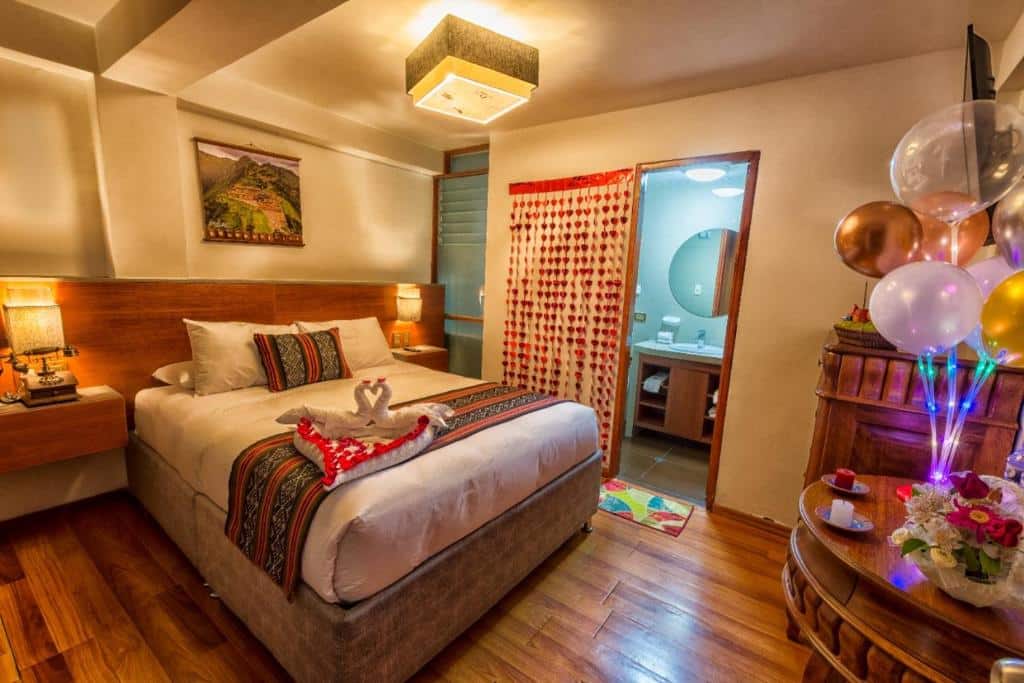 suíte romântica do chakana boutique hotel com uma cama de casal enfeitada com toalhas ornamentadas em formato de cisnes, com vários corações pendurados numa cortina ao lado da entrada do banheiro. Em frente a cama há uma mesa com vários balões coloridos e luzes divertidas decoradas em cima.