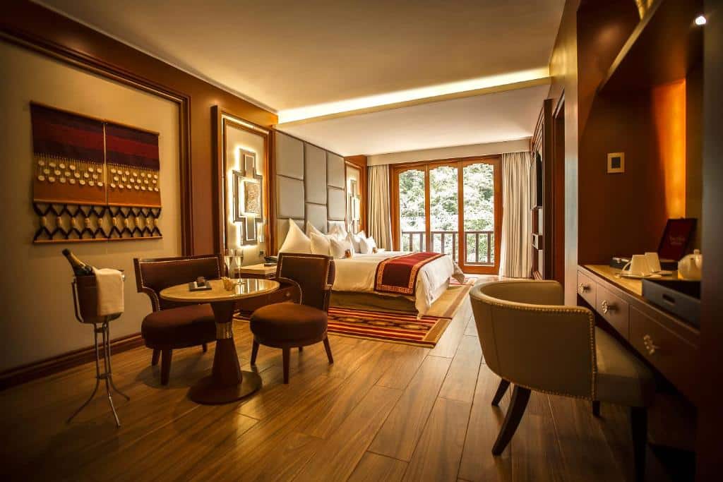 suíte master do hotel sumaq onde é possível ver uma mesa redonda pequena no lado esquerdo, acomodada com duas pequenas poltronas estofadas. Atrás da mesa há uma grande cama de casal, e atrás da cama, uma porta de vidro bem ampla com acesso a varanda privativa e vista para a paisagem natva.