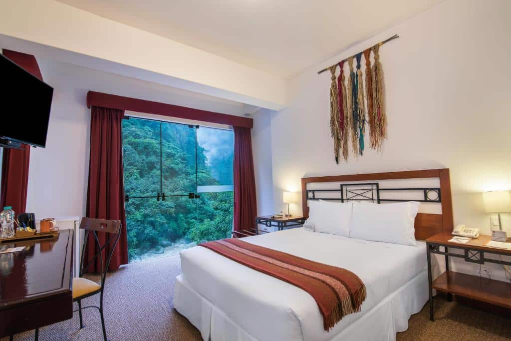 quarto do tierra viva machu picchu hotel com uma cama de casal no lado direito, uma mesa com cadeira em frente a cama e uma janela grande ao fundo que mostra a montanha atrás.