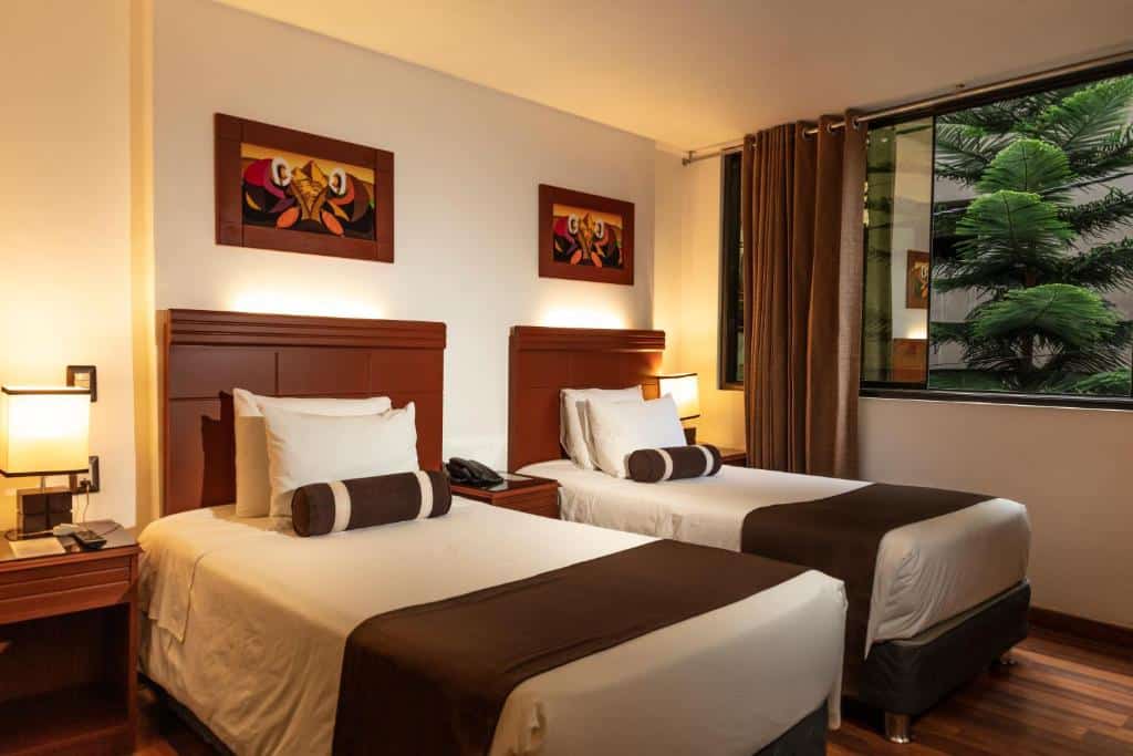 quarto duplo do hotel ferré boulevard com duas camas de solteiro, duas cômodas de cada lado das camas com abajures e uma janela com vista para a vegetação no canto direito superior da imagem.