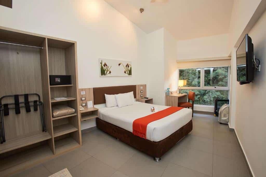 suíte da casa andina standard machu picchu com uma cama de casal, um armário sem portas com cabideiro, cofre e cobertores no lado esquerdo da imagem, e uma mesa com cadeira embaixo de uma ampla janela com vista para a vegetação no lado direito.