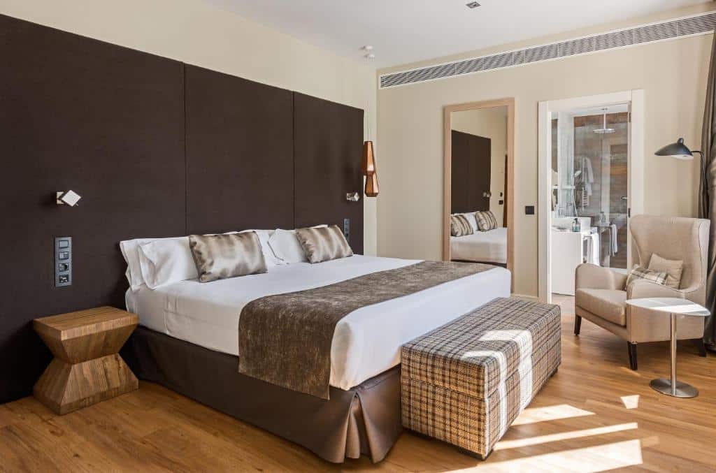 Quarto do Vincci The Mint, uma das recomendações de hotéis em Madri. Uma cama de casal com jogo de cama branco e mesinhas de cabeceira com luminárias dos dois lados encara uma TV na parede em frente. Uma mesinha com poltrona está no canto da parede que tem uma janela com cortinas verde e brancas. As paredes do quarto são ciano escuro.