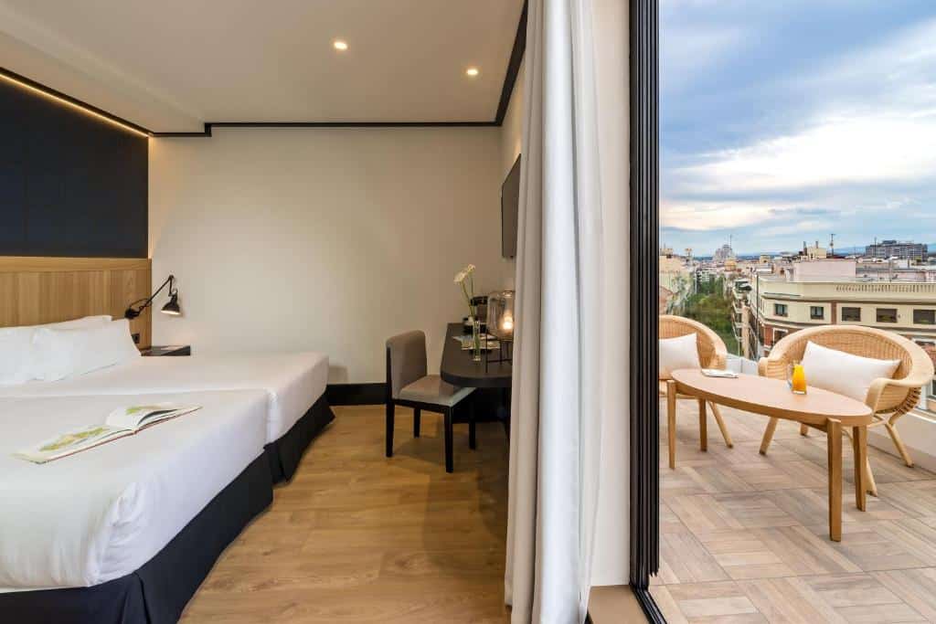 Vista dividida de um quarto do H10 Puerta de Alcalá, um dos hotéis em Madri. Duas camas com mesinha de cabeceira e abajur encaram uma mesa que tem cadeira, vaso de flores, abajur e uma TV na parede acima. Do outro lado, a varanda com mesa e cadeiras tem vista para a cidade. A porta que separa os ambientes tem cortinas bege.