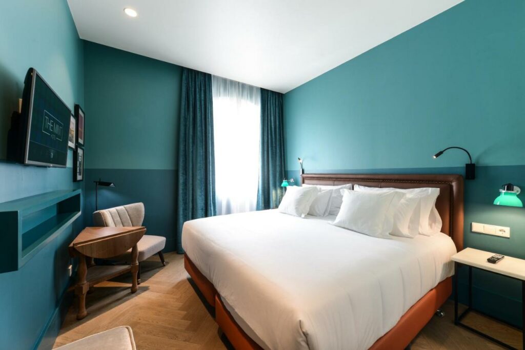 Quarto do Vincci The Mint, uma das recomendações de hotéis em Madri. Uma cama de casal com jogo de cama branco e mesinhas de cabeceira com luminárias dos dois lados encara uma TV na parede em frente. Uma mesinha com poltrona está no canto da parede que tem uma janela com cortinas verde e brancas. As paredes do quarto são ciano escuro.