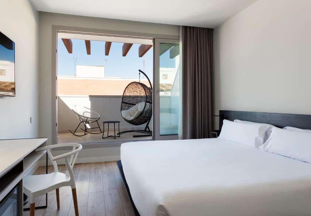 Quarto do B&B Hotel Madrid Centro Puerta del Sol, uma das recomendações de hotéis em Madri. A cama de casal está encostada na parede do lado esquerdo e encara uma mesa de trabalho, que tem cadeira e uma TV na parede acima. Ao fundo, uma grande porta de vidro que tem cortinas no canto está aberta e leva a um terraço com belas poltronas.