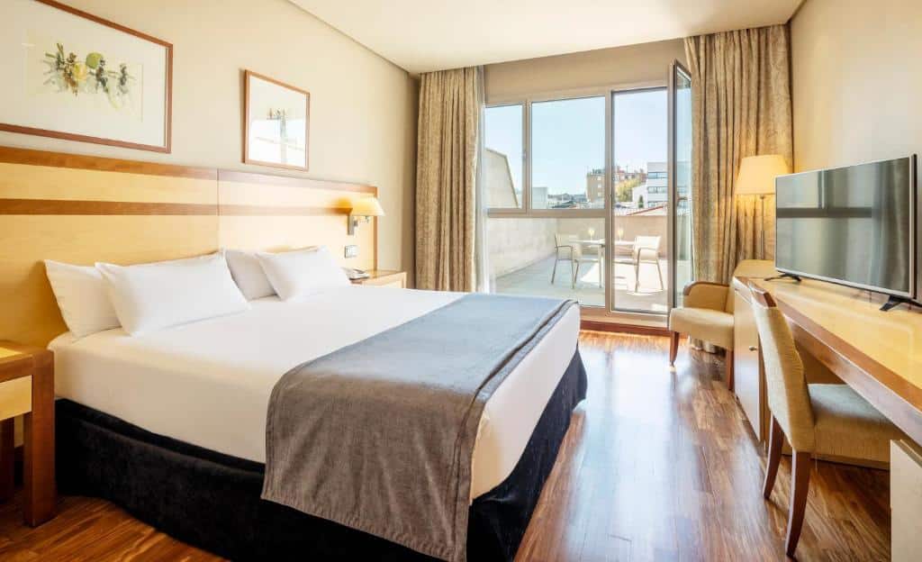 Quarto do Ilunion Alcalá Norte, uma das recomendações de hotéis em Madri. Uma cama de casal tem mesinhas de cabeceira e luminárias dos dois lados e encara uma mesa de trabalho com cadeira e TV. Ao fundo uma poltrona e um abajur estão em frente à porta de vidro que ocupa uma parede e leva à varanda com mesinha e cadeiras.