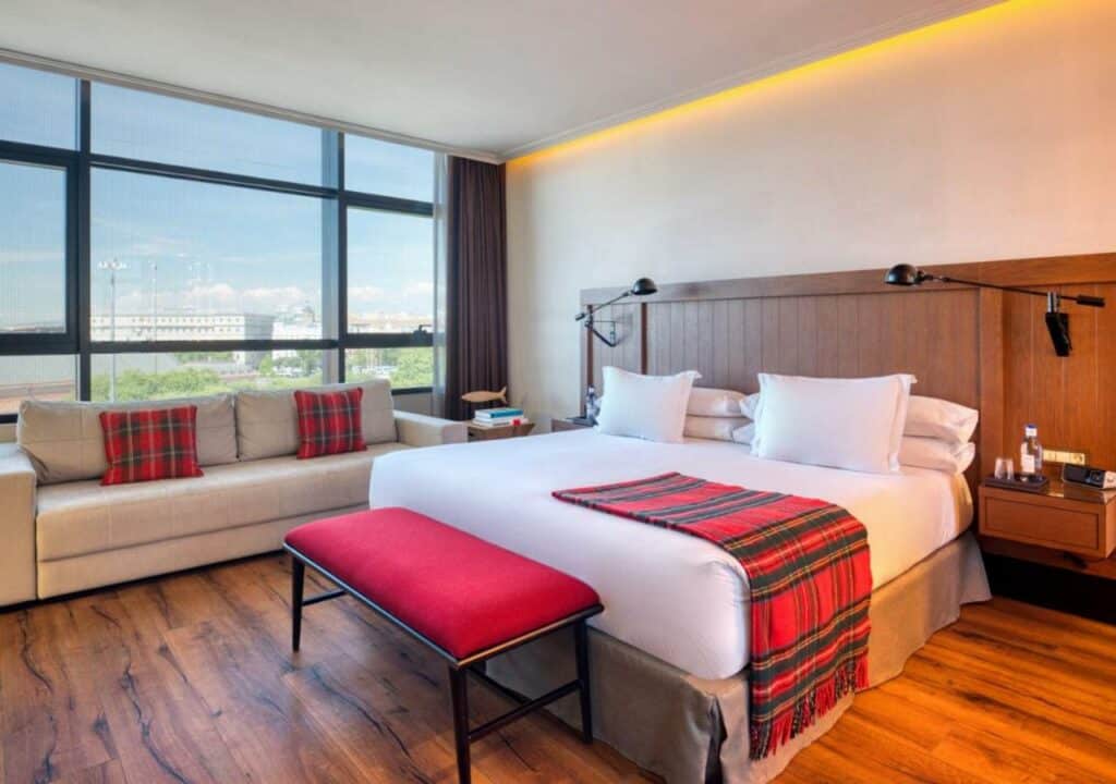 Quarto do Only YOU Hotel Atocha, uma das recomendações de hotéis em Madri. A cama de casal é ampla e tem um cobertor em cima e um banquinho aos pés. A cabeceira de madeira tem mesinhas com luminária dos dois lados. Há um sofá em frente da janela que ocupa a parede ao lado da cama. O piso é de madeira.