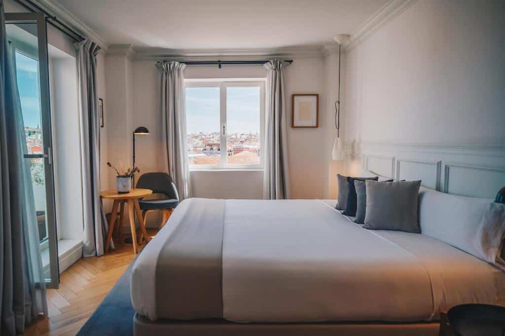 Quarto do Dear Hotel Madrid, uma das recomendações de hotéis em Madri. Uma cama de casal com jogo de cama branco e cinza encara uma porta de vidro aberta com cortinas cinza dos lados. Ao fundo, uma janela de vidro com cortinas cinza tem vista para a cidade, e há uma mesinha com vaso de plantas, luminária e cadeira ao lado esquerdo. Do lado direito, um quadrinho está pendurado na parede.