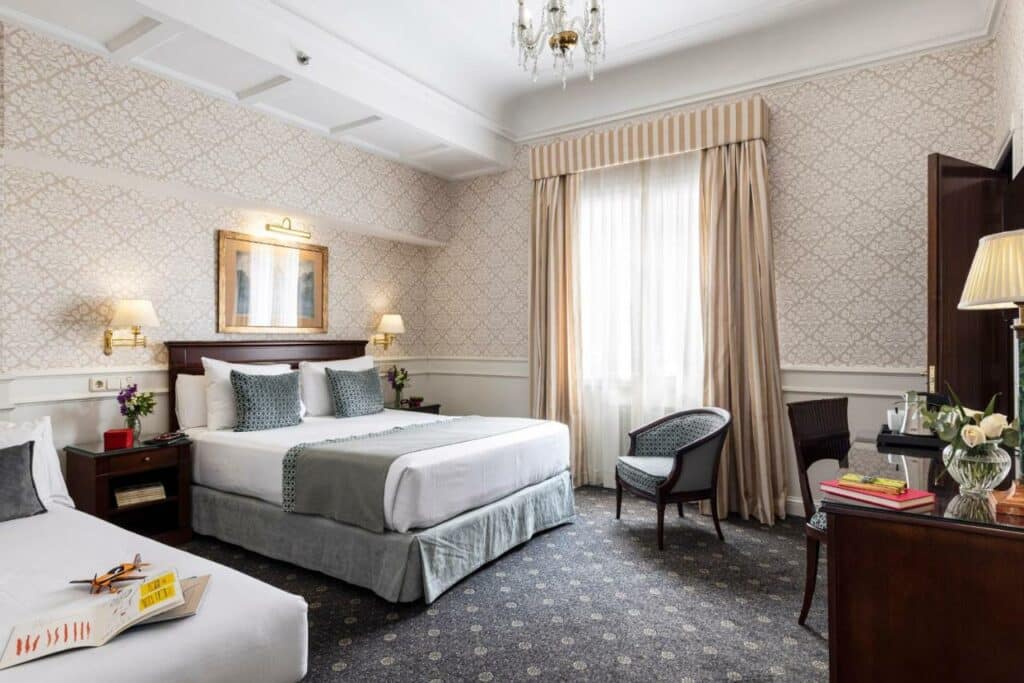 Quarto do Emperador, uma das recomendações de hotéis em Madri. Uma cama de casal tem mesinhas de cabeceira dos dois lados com vasos de flores e abajures, e encara uma mesa de trabalho com cadeira, abajur e comodidades para fazer chá. Há outra cama do lado esquerdo da de casal, e uma grande janela coberta com cortinas brancas está do lado direito. Há uma poltrona em frente à janela e perto da cama de casal.