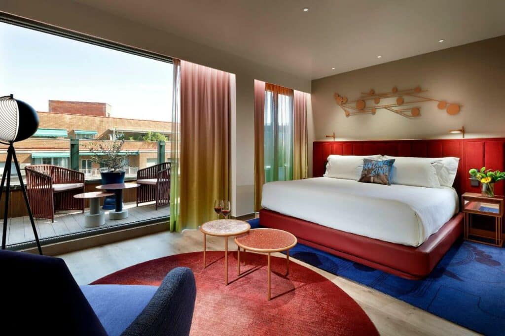 Quarto do Hard Rock Hotel Madrid, um dos hotéis em Madri. Uma ampla cama de casal está ao fundo do quarto e tem mesinhas de cabeceira e luminárias dos dois verdes. Uma área de estar em frente à cama conta com mesinha, poltrona, abajur e tapete. A parede ao lado tem amplas janelas e uma porta de vidro que leva à varanda com mesinha e cadeiras.