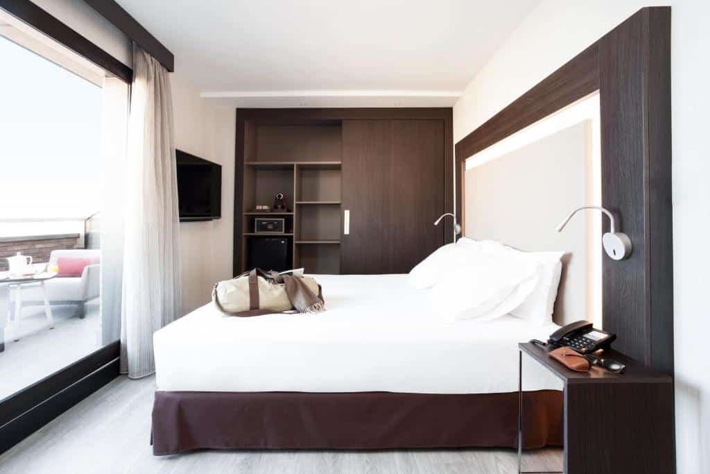 Quarto do Novotel Madrid Center, um dos hotéis em Madri. A cama de casal está encostada na parede ao lado direito e tem mesinhas de cabeceira e abajur dos dois lados. O guarda-roupa está no fundo do quarto, e uma TV na parede ao lado encara a cama. Uma porta de vidro em frente à cama tem cortinas brancas e vista para o terraço com poltronas e mesinha.
