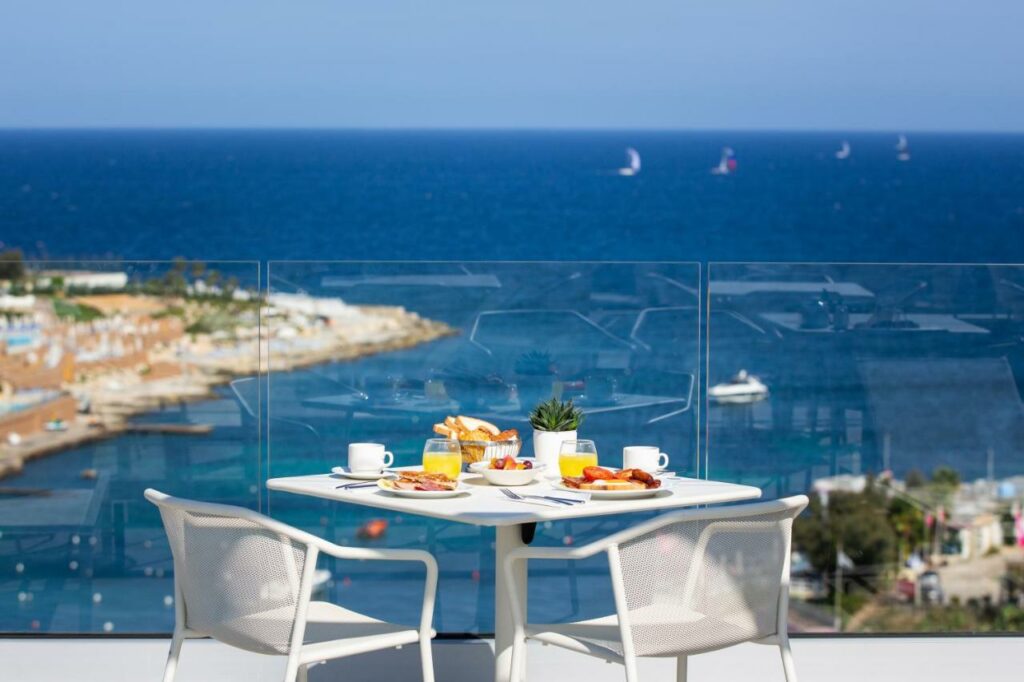 Varanda com vista para o mar no be.HOTEL com uma mesa e duas cadeiras. Há itens de café da manhã servidos sob a mesa. E placas transparentes que permitem ver o mar enquanto se está sentado.