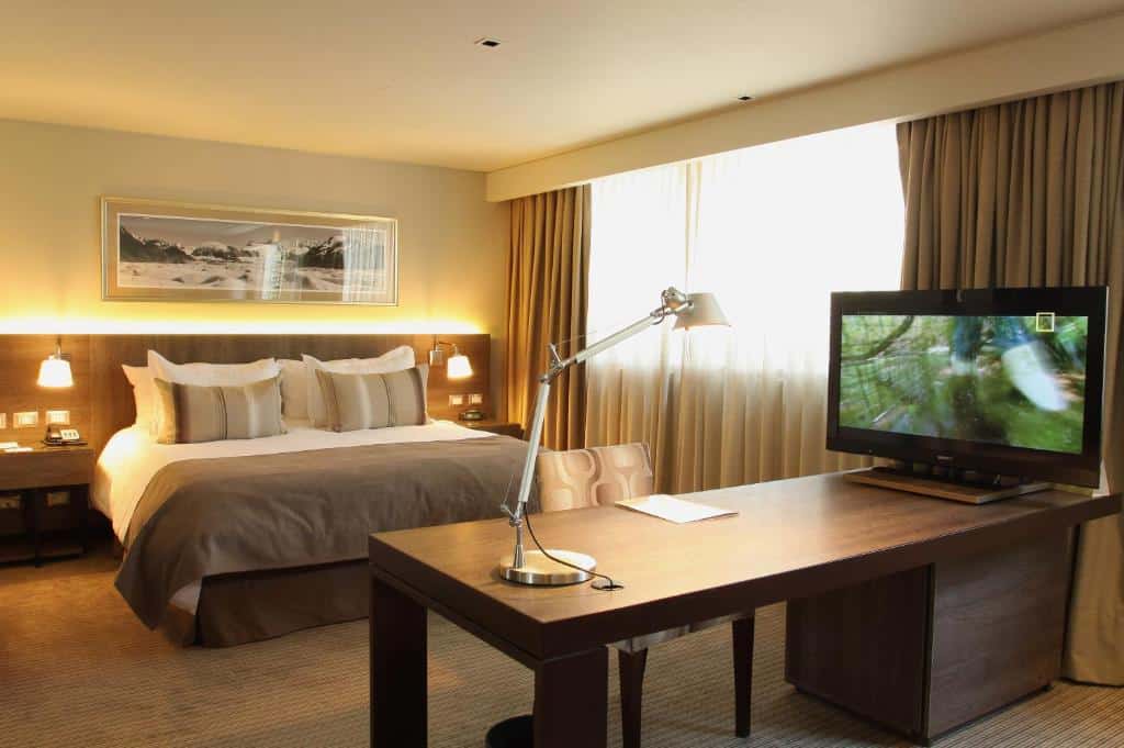 Quarto do Hotel Los Españoles Plus com cama de casal do lado esquerdo do quarto em frente cama uma mesa de trabalho com TV em cima. Representa hotéis em Providencia em Santiago.
