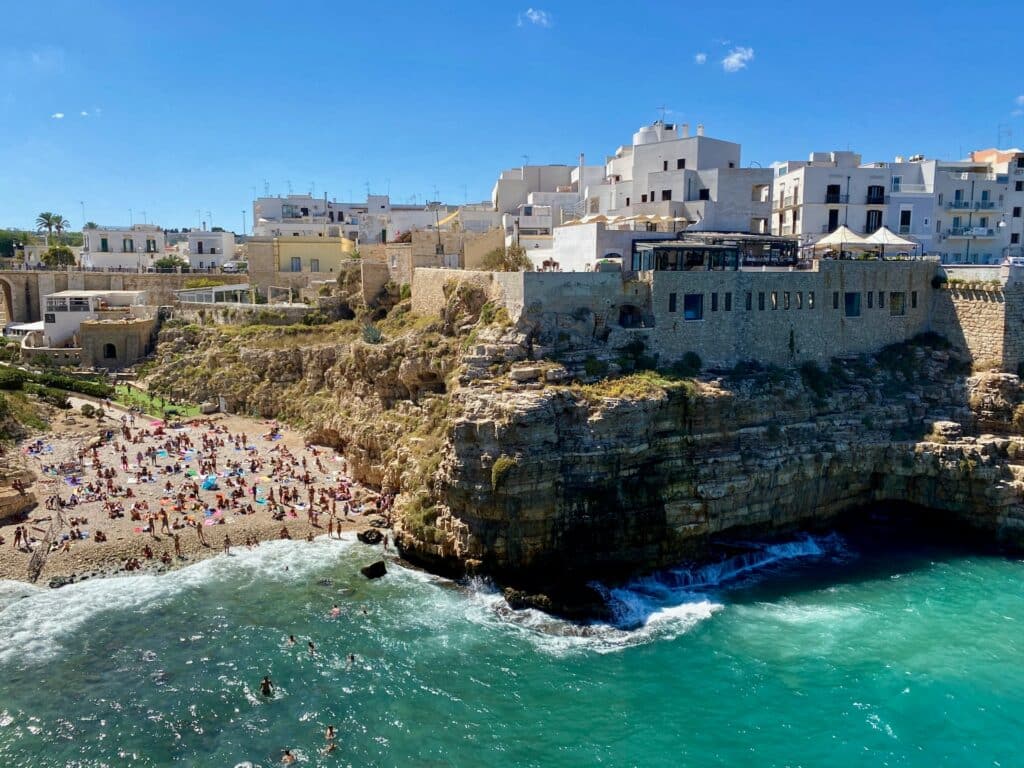Imagem aérea da pequena praia a esquerda com várias pessoas na faixa de areia e algumas na água azul e verde, em Polignano a Mare, em Puglia. Já do lado direito um paredão de pedra com várias casas em cima durante o dia, ilustrando post Hotéis em Puglia.