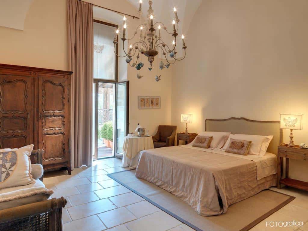 Quarto do Dimora Storica Torre Del Parco 1419 com uma cama de casal ao centro em cima de um tapete, ao lado direito tem uma mesinha com um abajur e do lado esquerdo tem outra mesinha com um abajur e uma poltrona com uma mesa redonda. Além disso, tem um sofá no quarto, um guarda-roupa de madeira, um lustre antigo e uma porta de vidro com cortinas.