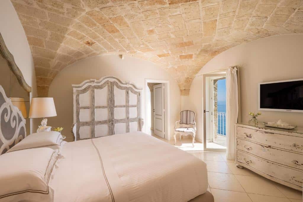 Quarto do Hotel Don Ferrante com uma cama de casal com dois travesseiros e uma mesinha com um abajur do lado. Na frente da cama tem uma cômoda com uma tv em cima, já do lado esquerdo do quarto tem um guarda-roupa, uma porta aberta que dá para outro cômodo e uma cadeira. Do lado da cadeira tem uma porta aberta que dá acesso a varanda do quarto.