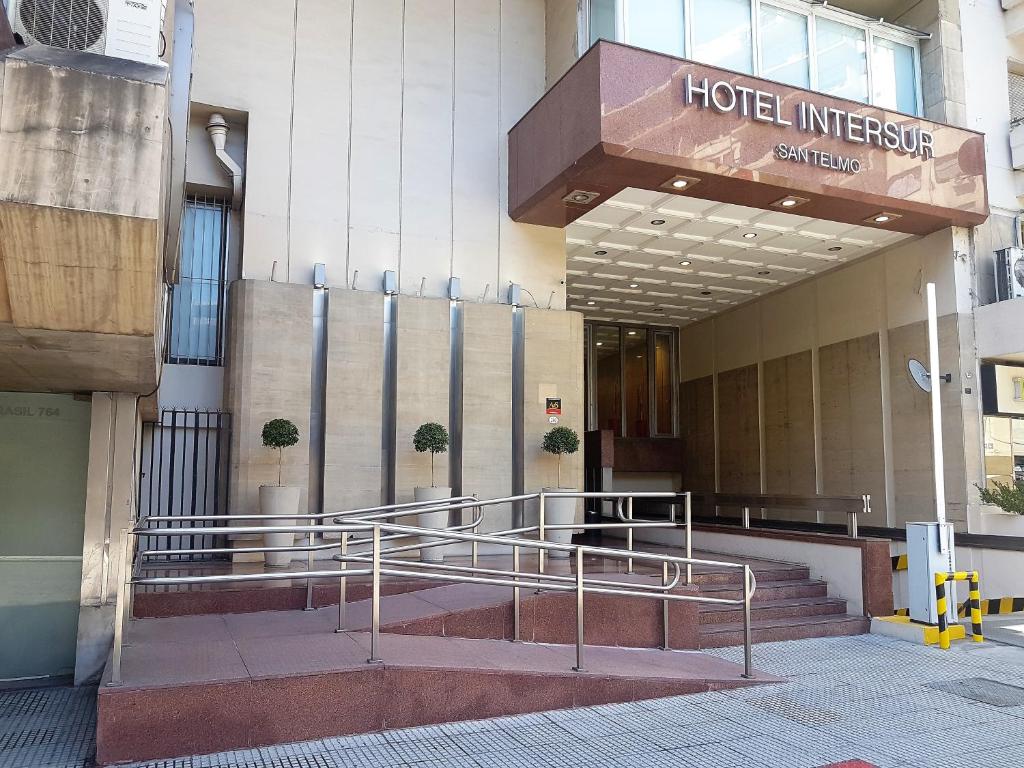 A imagem mostra a entrada do Hotel Intersur San Telmo em Buenos Aires, destacando uma fachada moderna com linhas retas e cores neutras. A entrada possui uma rampa de acesso acessível, com corrimãos de metal, facilitando a mobilidade para pessoas com necessidades especiais. Ao lado da rampa, há escadas que levam até a porta principal. A fachada é adornada com pequenas árvores em vasos, que adicionam um toque de verde ao ambiente. A cobertura da entrada exibe o nome do hotel em letras grandes e claras.