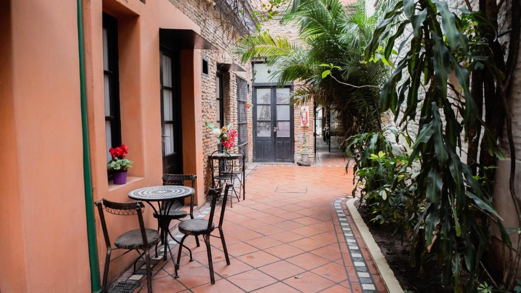 A imagem mostra a área comum do Pasaje Solar, um dos hotéis em San Telmo, Buenos Aires. Há um jardim interno com plantas altas e bem verdes, é possível ver ainda portas de vidro com estrutura de metal, no mesmo tom escuro das mesinhas com cadeiras de ferro do lado de fora. O chão é em tons terrosos.