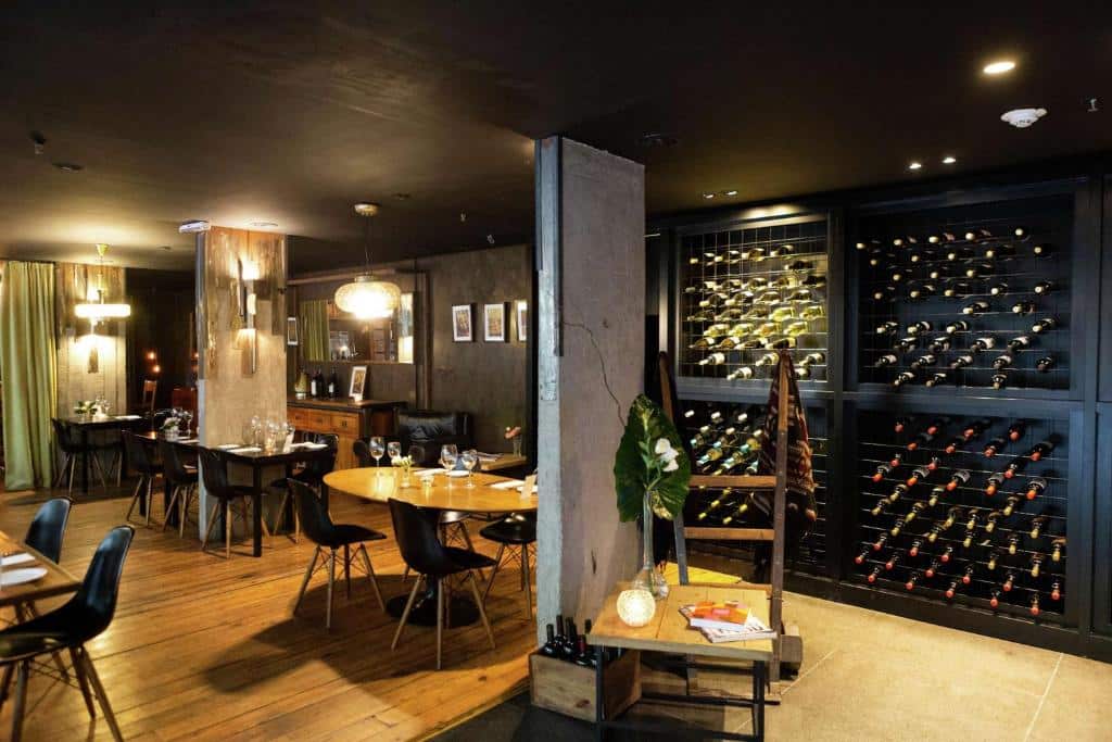 A imagem mostra o lounge do Anselmo Buenos Aires, Curio Collection by Hilton, decorado com um ambiente sofisticado e aconchegante. Há várias mesas de jantar dispostas em um espaço com piso de madeira, cercadas por cadeiras de design moderno. À esquerda, luminárias pendentes proporcionam uma iluminação suave e acolhedora. À direita, uma impressionante adega ocupa uma parede inteira, exibindo uma vasta seleção de vinhos. O ambiente combina elementos rústicos com um toque contemporâneo, criando um espaço elegante e convidativo para refeições ou degustação de vinhos.