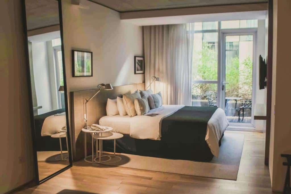A imagem mostra um quarto do Anselmo Buenos Aires, Curio Collection by Hilton, decorado com um estilo moderno e acolhedor. A cama é ampla e está equipada com várias almofadas, proporcionando conforto. À esquerda, um espelho de corpo inteiro amplia a sensação de espaço, enquanto uma luminária de leitura está posicionada ao lado da cama sobre uma mesa de apoio. Ao fundo, grandes janelas com cortinas transparentes permitem a entrada de luz natural, criando um ambiente iluminado e agradável. Através das janelas, pode-se ver uma pequena varanda com uma mesa e cadeiras, oferecendo um espaço adicional para relaxamento. O piso de madeira clara contribui para a atmosfera acolhedora do quarto.