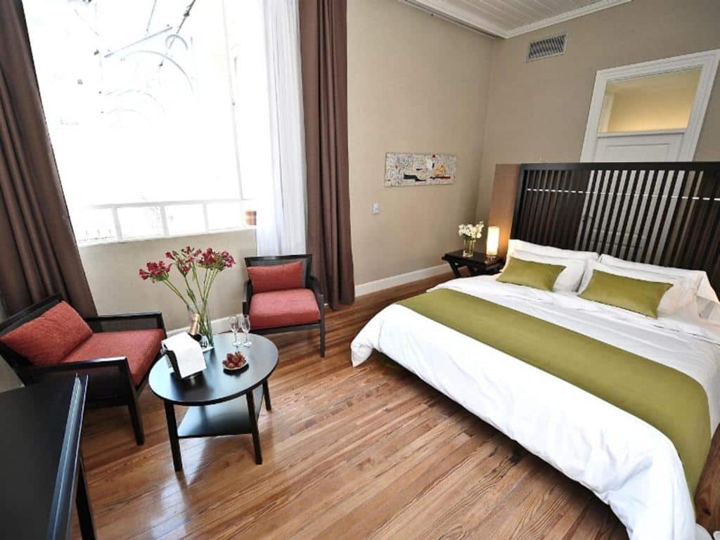 A imagem mostra um quarto elegante no Patios de San Telmo, com uma cama de casal espaçosa decorada com almofadas e peseira verdes. Há uma área de estar com duas poltronas acolchoadas em cor laranja e uma mesa de centro preta, onde estão dispostos uma garrafa de champanhe e copos. O piso de madeira clara e as cortinas longas que enquadram a janela criam um ambiente acolhedor e iluminado.