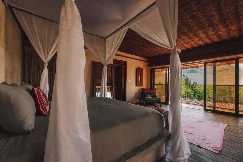 Suíte Júnior deluxe do Radhoo Tulum, de 53 m², com cama de casal, espelho ao lado, poltrona azul à frente perto da parede e porta extensa de vidro que dá acesso à varanda, que tem uma visão panorâmica da natureza, para representar hotéis em Tulum.