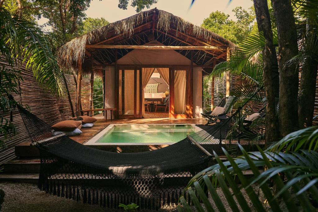 Uma villa com telhado de palha com piscina externa, rede e espreguiçadeiras cercadas por vegetação e árvores exuberantes, no Habitas Tulum, para representar hotéis em Tulum.