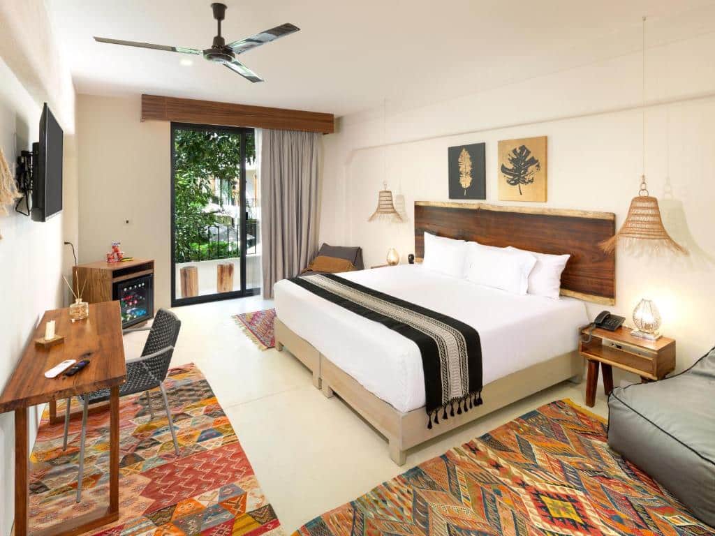 Quarto de um apartamento do Naala Tulum, de 110 m², sendo dentro do quarto uma cama de casal, uma mesinha com uma cadeira, uma tv suspensa na parede, escrivaninha ao lado da cama, poltrona cinza e uma pequena varanda com vista para árvores e outras acomodações do local, para representar hotéis em Tulum.