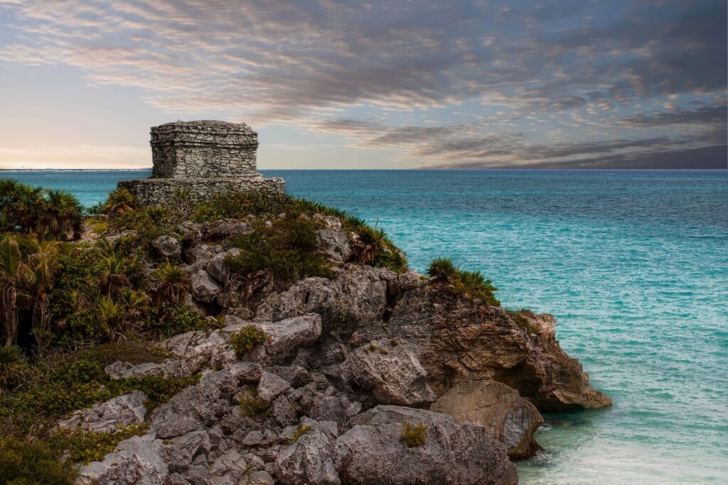 Área costeira do mar na Praia de Tulum, com uma montanha de rochas, uma construção antiga ao fundo, e o mar cristalino azul ao lado.