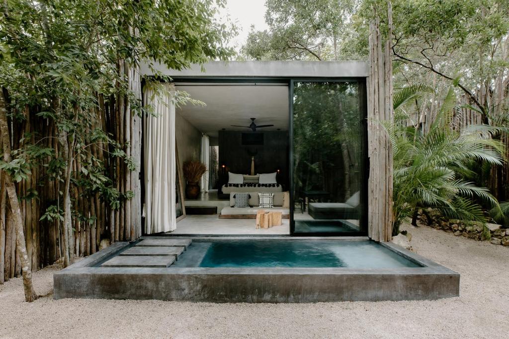 Uma villa moderna com grandes portas de vidro, uma pequena piscina privativa e assentos ao ar livre, cercada por árvores e vegetação, no Hotel Bardo, para representar hotéis em Tulum.