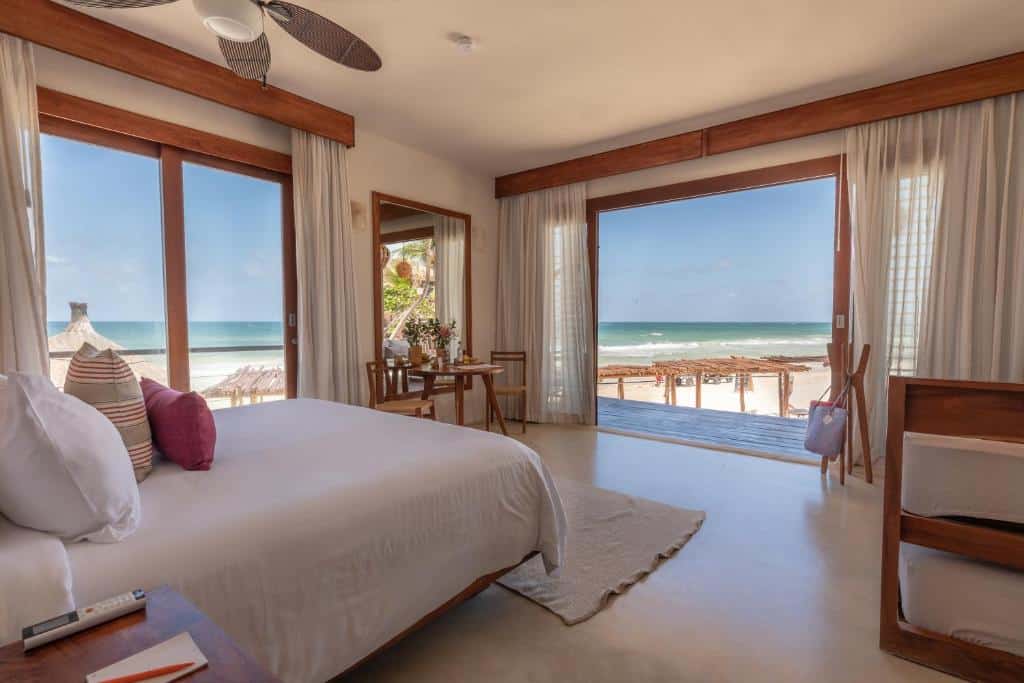 Suíte à beira-mar do La Zebra a Colibri Boutique Hotel, de 40 m², com cama de casal, bicama de solteiro no lado direito, mesa redonda com flores em cima e cadeiras em volta, além de duas portas grandes de vidro com vista para a praia, para representar hotéis em Tulum.