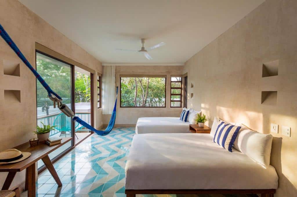 Quarto do Tiki Tiki Tulum, de 30 m², com duas camas de casal, rede azul pendurada de ponta à ponta na frente das camas, mesa com vaso de flor e chapeu em cima, e uma varanda com cadeira azul e vista para a natureza, para representar hotéis em Tulum.