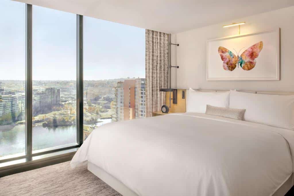 Quarto no JW Marriott Parq Vancouver com uma ampla janela que vai do texto até o chão dando vista para uma baía e outro prédios, dentro do quarto há uma cama de casal e sob ela há um quadro com uma borboleta desenhada, as paredes são brancas e o piso tem carpete em tom de bege.