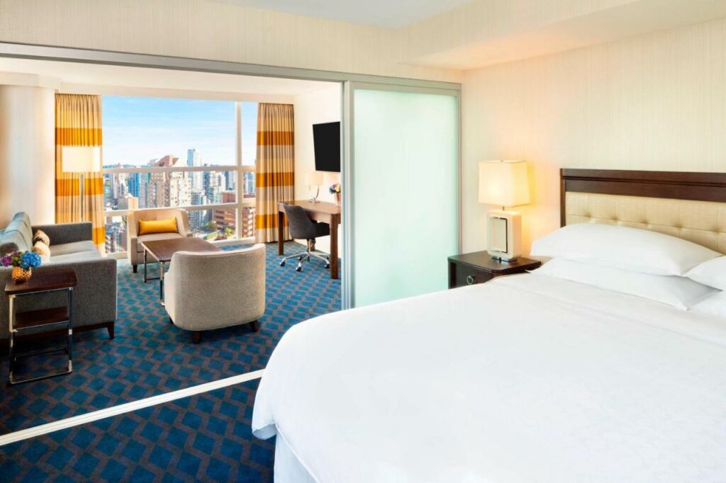 Quarto do Sheraton Vancouver Wall Centre com ambiente duplo, do lado esquerdo há uma sala de estar com sofá, poltronas, televisão e uma ampla janela com cortinas, do outro lado, uma cama de casal, há carpete azul com detalhes cinza em todo o ambiente, para representar hotéis em Vancouver.