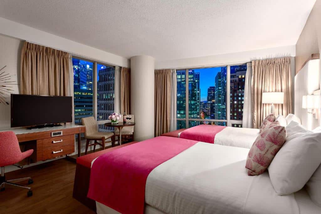 Quarto do Auberge Vancouver Hotel com janela panorâmica com vista para a cidade, há uma pequena mesinha com duas cadeiras perto de uma das janelas, ao lado, uma mesa de escritório com uma televisão e uma cadeira giratória, do lado direito, há duas camas de solteiro com travesseiros e almofadas, o chão é de madeira.